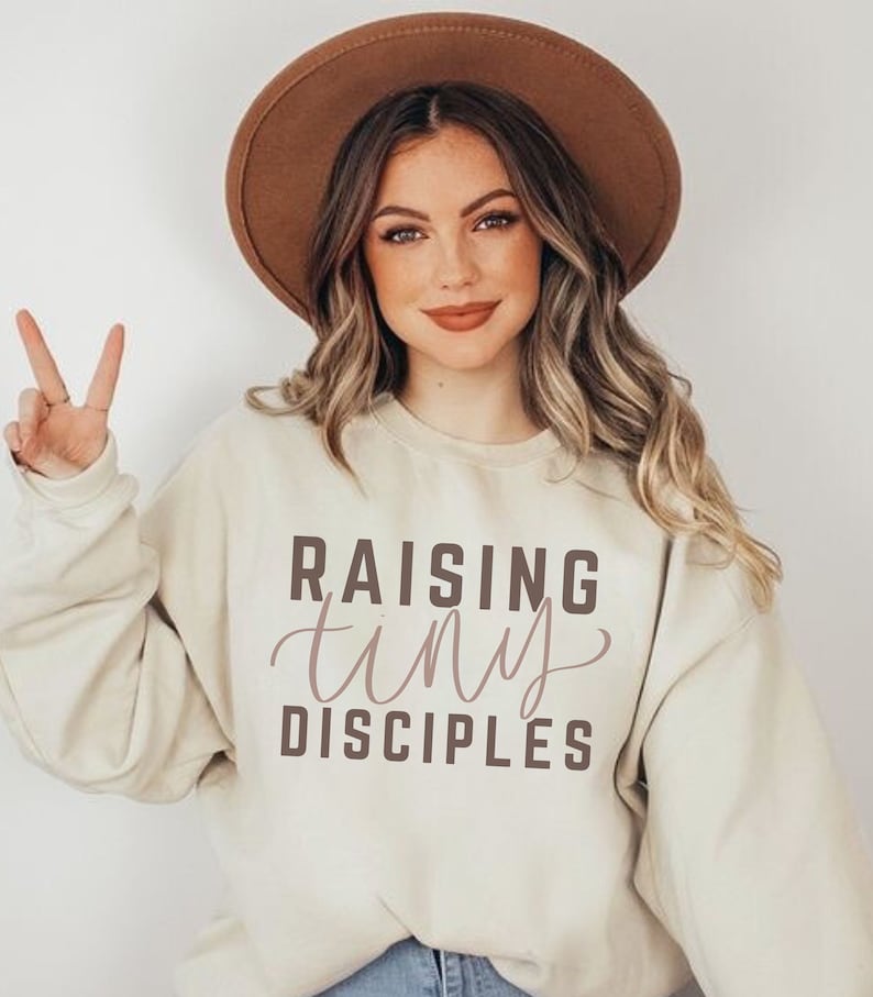 May include: A beige sweatshirt with the text "Raising tiny disciples" printed in brown on the front.