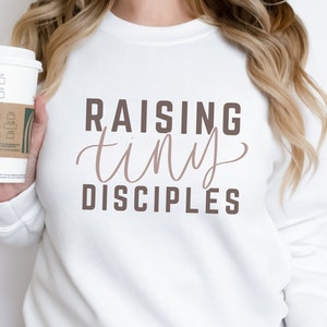 May include: White sweatshirt with the text "Raising tiny disciples" in brown lettering. The word "tiny" is in a cursive font.