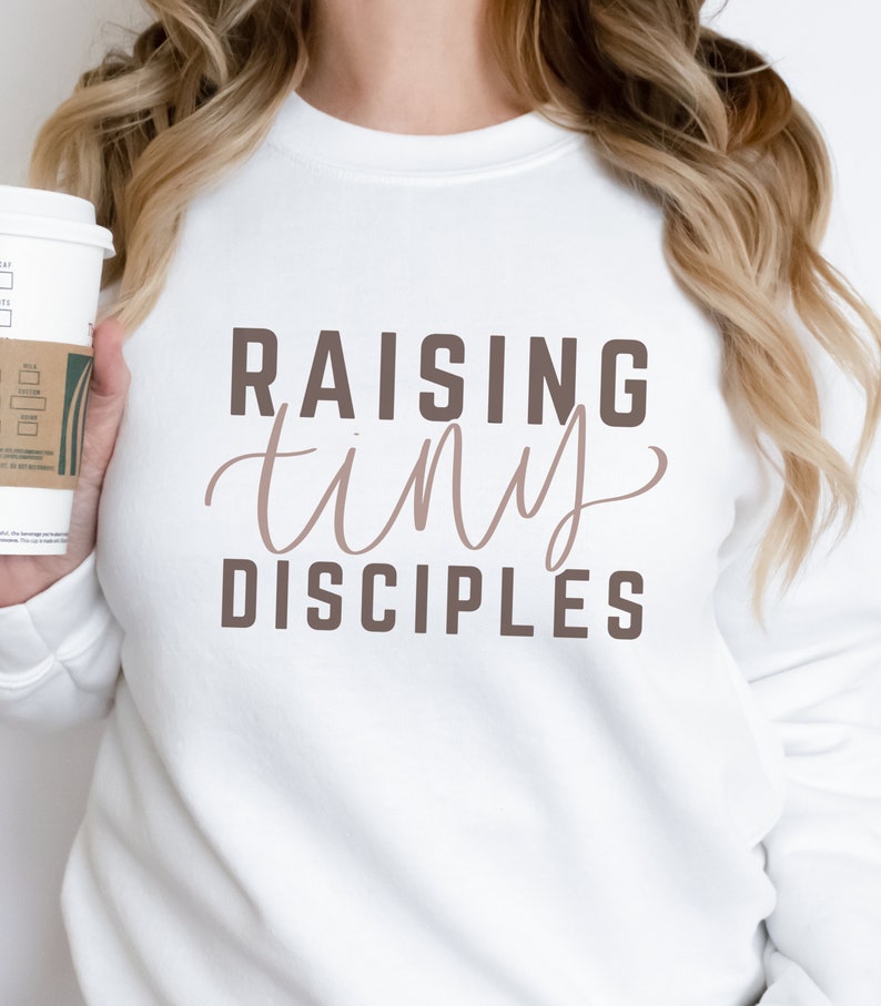 May include: White sweatshirt with the text "Raising tiny disciples" in brown lettering. The word "tiny" is in a cursive font.