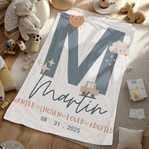 May include: White baby blanket with a large blue letter "M" and the name "Martin" in cursive. The blanket features a sun, stars, a car, and a cloud with hearts. The words "WANTED, CHOSEN, LOVED, ADOPTED" and the date "08.31.2025" are also printed.