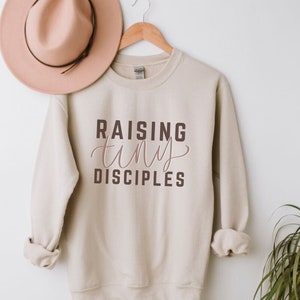 May include: A beige sweatshirt with the text "RAISING tiny DISCIPLES" in brown and pink lettering.