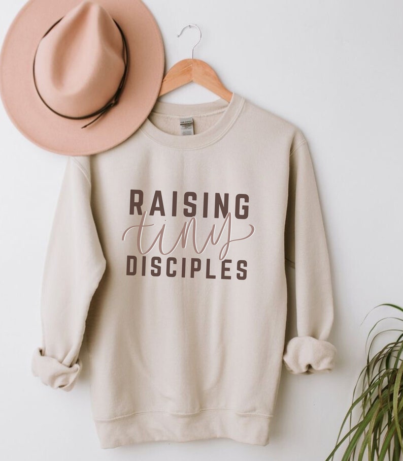 May include: A beige sweatshirt with the text "RAISING tiny DISCIPLES" in brown and pink lettering.