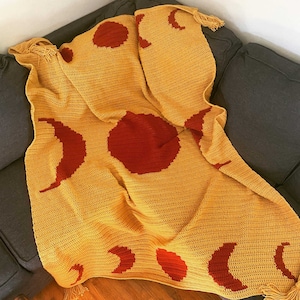 May include: A crocheted throw blanket in a mustard yellow color with a repeating pattern of red moons and suns. The blanket has fringe on the edges.