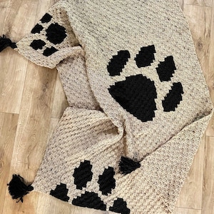 Puede incluir: Una manta de punto de ganchillo beige con diseños de huellas de patas negras. La manta tiene borlas negras en cada esquina.