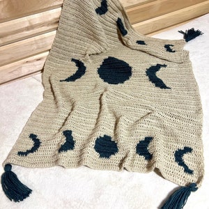 Può includere: Una coperta all'uncinetto con sfondo beige e disegni di luna e sole verde scuro. La coperta ha frange su ogni angolo.