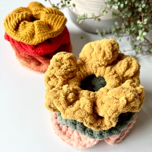 May include: A stack of four crocheted hair scrunchies in various colors, including mustard yellow, red, orange, and green. The scrunchies are made with a soft, fuzzy yarn.