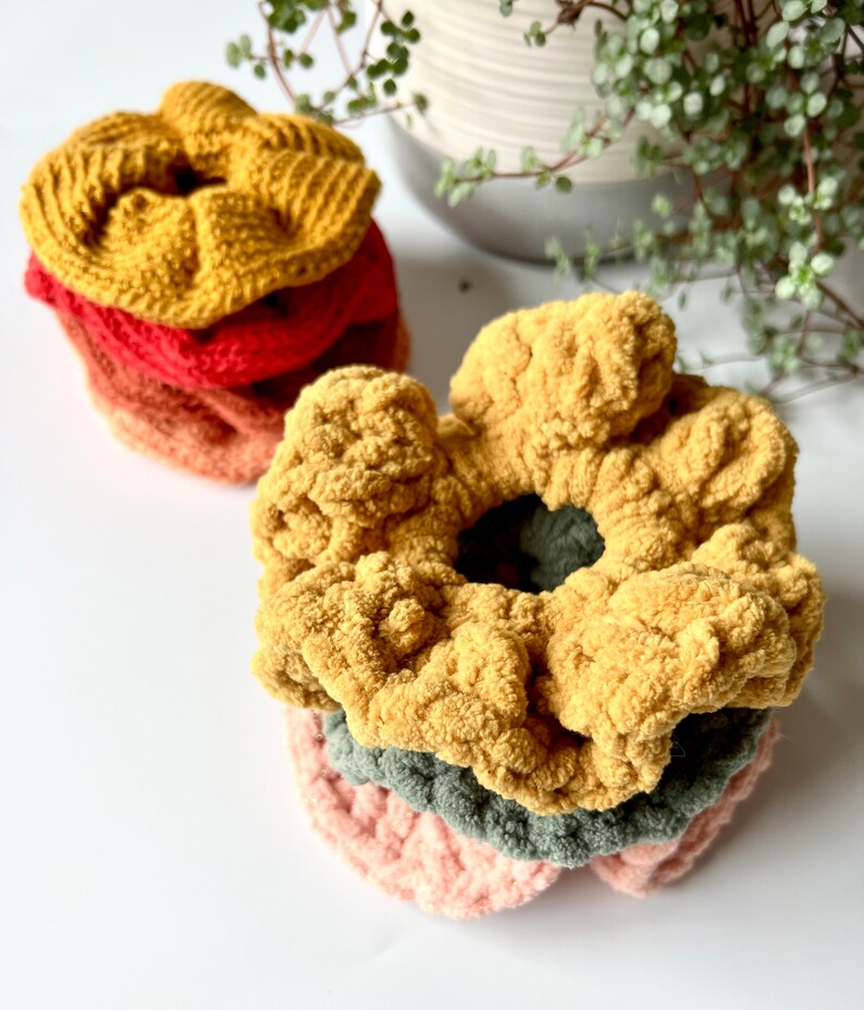 May include: A stack of four crocheted hair scrunchies in various colors, including mustard yellow, red, orange, and green. The scrunchies are made with a soft, fuzzy yarn.