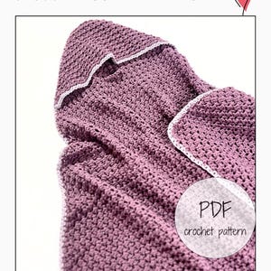 Puede incluir: Una toalla de bebé de color malva, tejida a ganchillo, con capucha y ribete blanco. La toalla está doblada, mostrando el patrón de puntadas texturizadas. El texto "SNUGGLE HOODED BABY TOWEL" está en la parte superior, y un gráfico "PDF crochet pattern" está en la esquina inferior derecha.