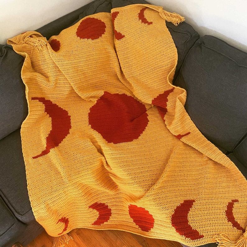 May include: A crocheted throw blanket in a mustard yellow colour with a repeating pattern of red moons and suns. The blanket has fringe on the edges.