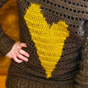 Pu&ograve; includere: Un maglione di uncinetto marrone con un grande disegno a forma di cuore giallo.