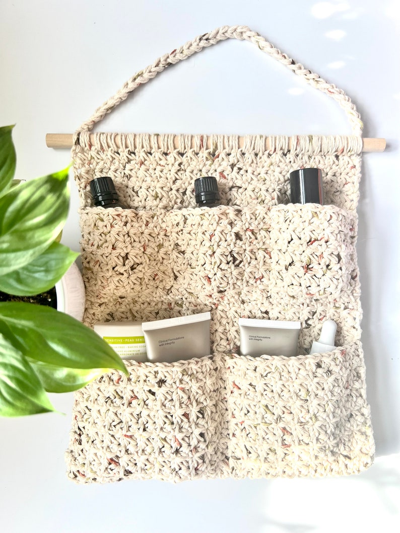 May include: A beige crocheted wall hanging with four pockets. The pockets contain three small bottles and three tubes of cream. The wall hanging has a wooden dowel rod at the top.