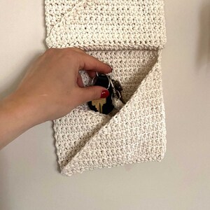 Pu&ograve; includere: Una tasca da parete in crochet bianco con una patta che si ripiega per creare una piccola tasca. Le chiavi sono riposte all'interno della tasca.