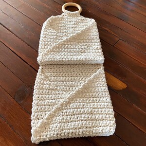 Pu&ograve; includere: Una borsa a tracolla in crochet beige con un manico ad anello in legno. La borsa &egrave; piegata a met&agrave;, mostrando il motivo a uncinetto.