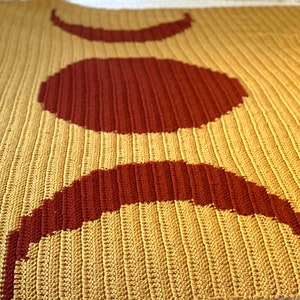 May include: A crocheted throw blanket featuring a design of three crescent moons in shades of red and yellow.