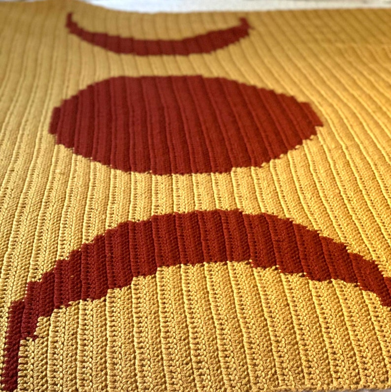 May include: A crocheted throw blanket featuring a design of three crescent moons in shades of red and yellow.