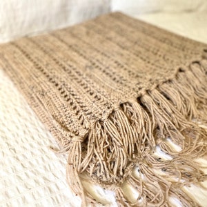 May include: A beige crocheted throw blanket with fringe. The blanket is made of a soft, textured yarn and has a simple, elegant design.
