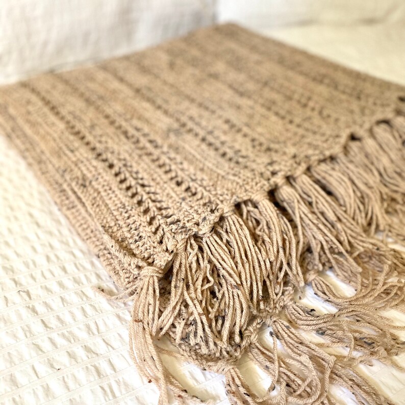 May include: A beige crocheted throw blanket with fringe. The blanket is made of a soft, textured yarn and has a simple, elegant design.