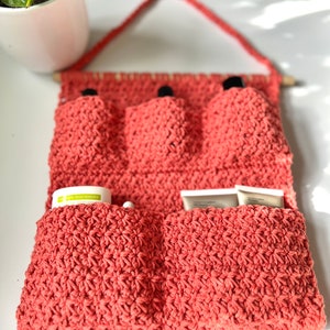 May include: A coral-colored crocheted wall organizer with three pockets for storing small items, such as makeup brushes or toiletries. The organizer has a wooden dowel at the top for hanging.