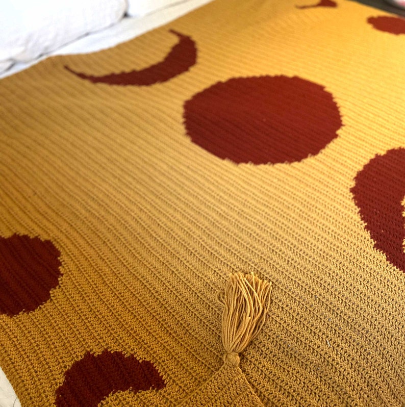 May include: A crocheted throw blanket with a mustard yellow background and red moon and sun designs. The blanket has a tassel on the bottom right corner.