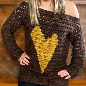 Pu&ograve; includere: Un maglione di uncinetto marrone con un grande disegno a forma di cuore giallo. Il maglione ha una vestibilit&agrave; ampia e una scollatura leggermente scesa.