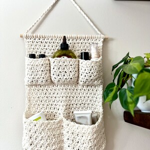 May include: A white crocheted wall hanging with six pockets for storing toiletries. The hanging has a wooden dowel at the top and is hung on a white wall. The pockets contain various bottles and tubes of toiletries.