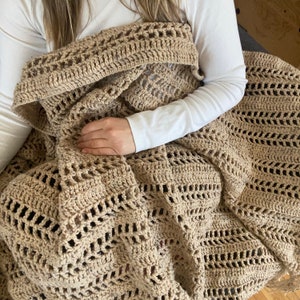 May include: A crocheted throw blanket in a beige color with a lacy pattern. The blanket is draped over a person sitting on a couch.