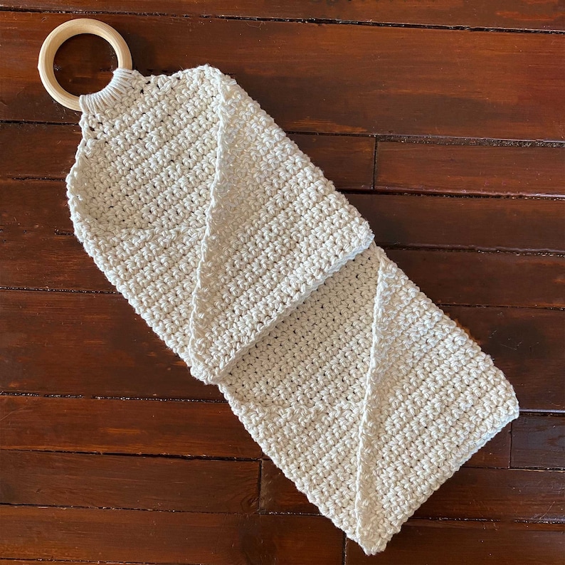 Pu&ograve; includere: Una borsa di stoccaggio sospesa in crochet beige con un anello di legno in cima.