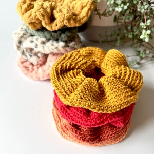 May include: A stack of four crocheted hair scrunchies in various colors, including yellow, pink, red, and orange. The scrunchies are made with a textured yarn and are stacked on top of each other.