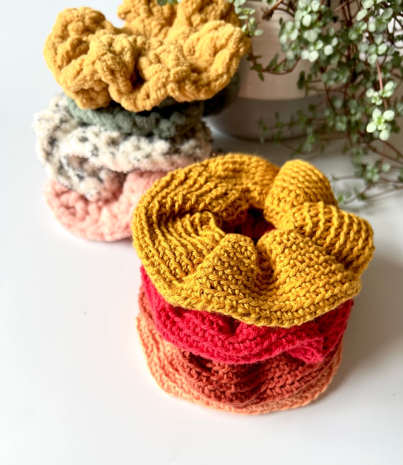 May include: A stack of four crocheted hair scrunchies in various colors, including yellow, pink, red, and orange. The scrunchies are made with a textured yarn and are stacked on top of each other.