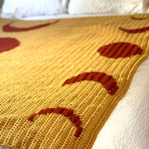 May include: A crocheted throw blanket in a mustard yellow colour with a pattern of red crescent moons.