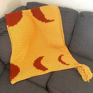 May include: A crocheted throw blanket in yellow with red crescent moon designs. The blanket has a fringe tassel on one end.