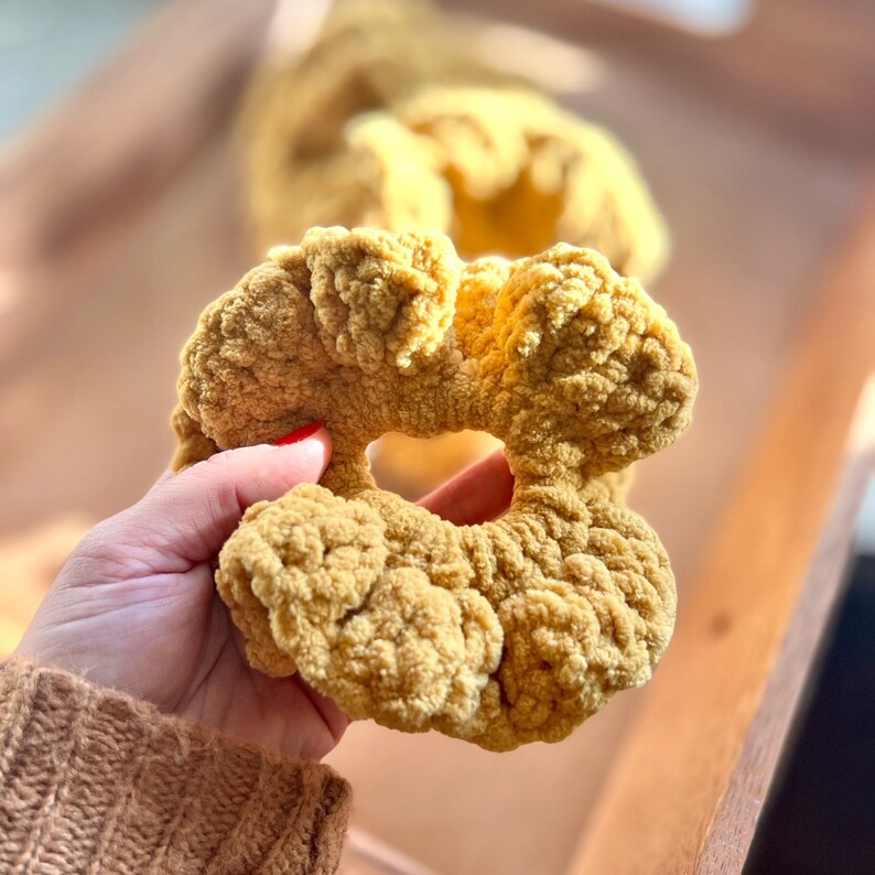 May include: A close-up of a crocheted flower-shaped hair scrunchie in a mustard yellow color.