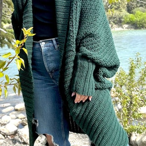 Pu&ograve; includere: Un cardigan di uncinetto verde scuro con maniche lunghe e una vestibilit&agrave; ampia. Il cardigan &egrave; aperto sul davanti e ha uno stile rilassato e casual.