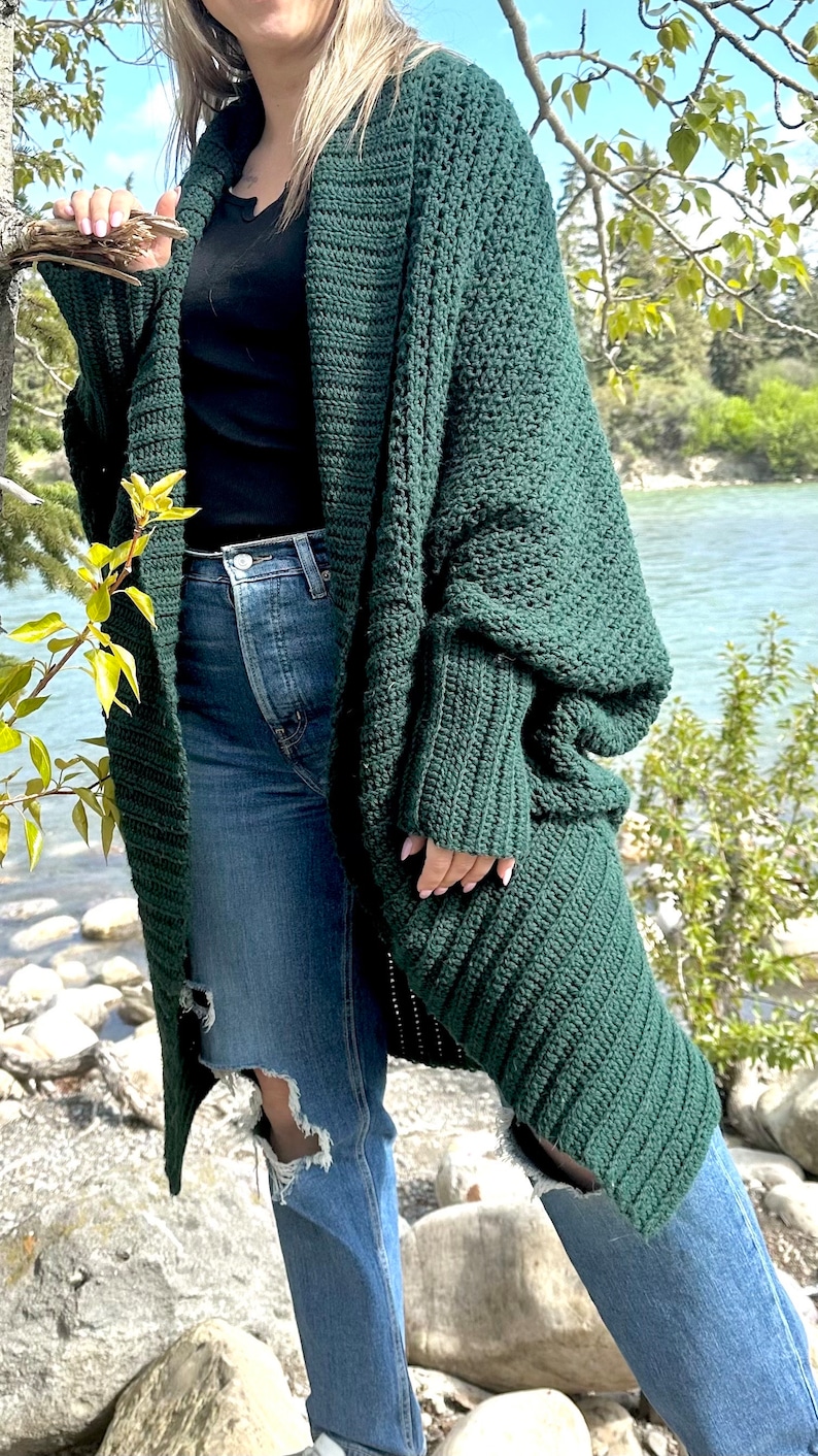 Pu&ograve; includere: Un cardigan di uncinetto verde scuro con maniche lunghe e una vestibilit&agrave; ampia. Il cardigan &egrave; aperto sul davanti e ha uno stile rilassato e casual.