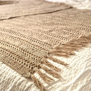May include: A beige crocheted throw blanket with fringe on the edges. The blanket is laying on a white bedspread.