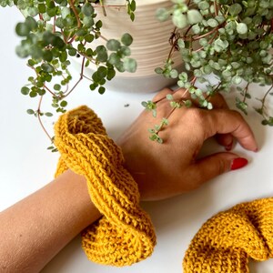 May include: Two mustard yellow knitted hair scrunchies. One is on a person's wrist, the other is lying on a white surface.