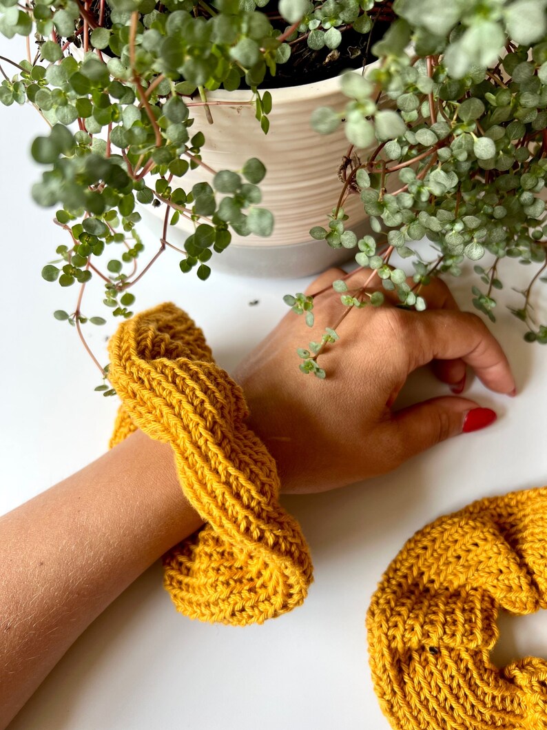 May include: Two mustard yellow knitted hair scrunchies. One is on a person's wrist, the other is lying on a white surface.