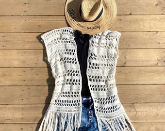 May include: White crochet vest with fringe detailing, worn over a black tank top and blue denim shorts. A straw hat with the text "Take me away..." is on top of the vest.