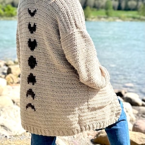 Puede incluir: Un cárdigan de crochet beige con un diseño de fases lunares negras en la espalda. El cárdigan es largo y tiene un corte holgado.