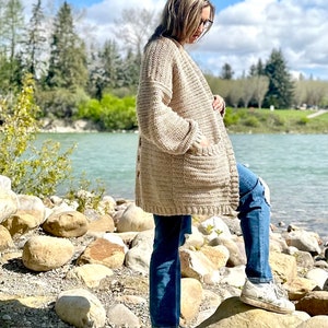 May include: A beige crocheted cardigan with long sleeves and a pocket on the front. The cardigan is worn by a person standing on a rocky shore.