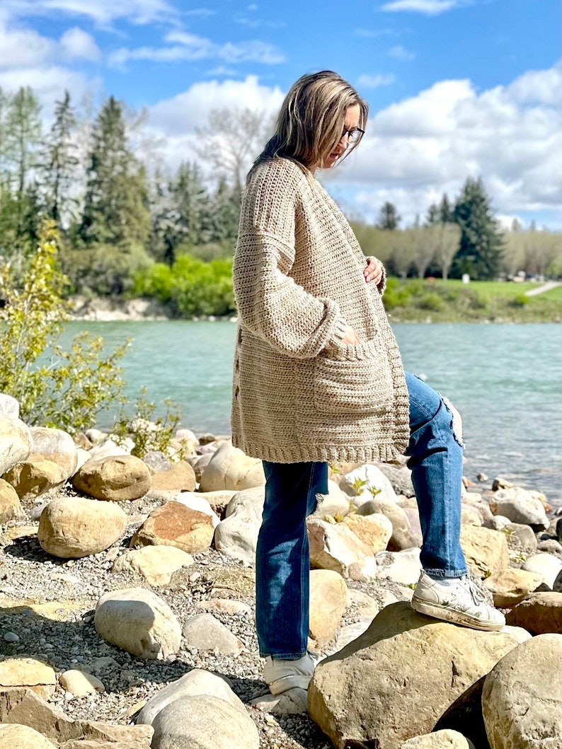 May include: A beige crocheted cardigan with long sleeves and a pocket on the front. The cardigan is worn by a person standing on a rocky shore.
