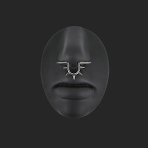 Op de afbeelding: Zilverkleurige neusring met een U-vormig ontwerp en meerdere spikes. De sieraden worden getoond op een zwart gezichtsmodel, waarbij de details en stijl van de piercing worden benadrukt. Een opvallend accessoire.