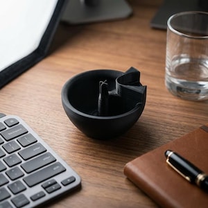 May include: A black, 3D-printed ashtray with a unique design, featuring a central post and a curved rest. The ashtray sits on a wooden desk, alongside a keyboard, a glass of water, a notebook, and a pen.