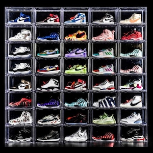 May include: A collection of trainers displayed in clear, stackable shoe storage boxes. The trainers feature a range of colours, such as blue, red, white, and green, with diverse designs and styles. The boxes are arranged in a grid against a black backdrop.