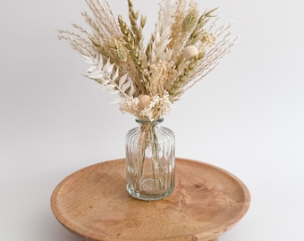 May include: A clear glass vase with a ribbed design holds a bouquet of dried flowers in shades of beige, cream, and green. The vase sits on a round, wooden tray with small, rounded feet. The arrangement is set against a white background.