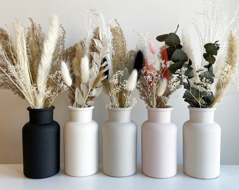 May include: Five ceramic vases in different colors, each filled with dried flowers and grasses. The vases are arranged in a row on a white surface.