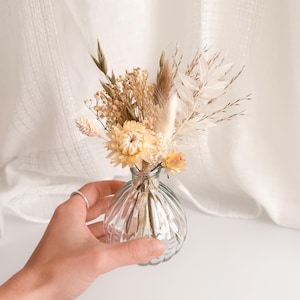 May include: A small, clear glass vase with a ribbed design, holding a bouquet of dried flowers. The flowers are in shades of cream, beige, and light yellow. The vase is held by a hand, and the background is a soft white fabric.