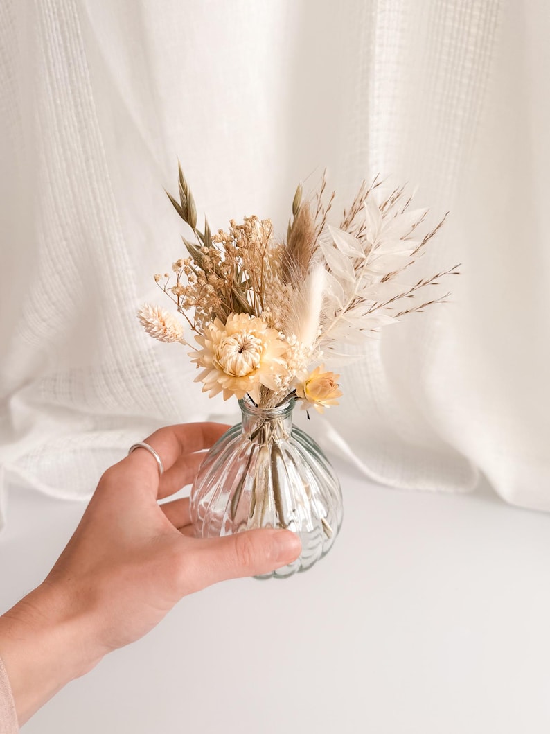 May include: A small, clear glass vase with a ribbed design, holding a bouquet of dried flowers. The flowers are in shades of cream, beige, and light yellow. The vase is held by a hand, and the background is a soft white fabric.