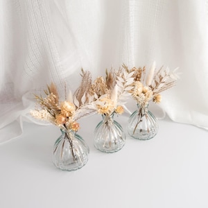 May include: Three small, clear glass vases with a ribbed design, each holding a delicate arrangement of dried flowers in shades of beige, cream, and light brown. The vases are arranged on a white surface, with a soft, white fabric backdrop.
