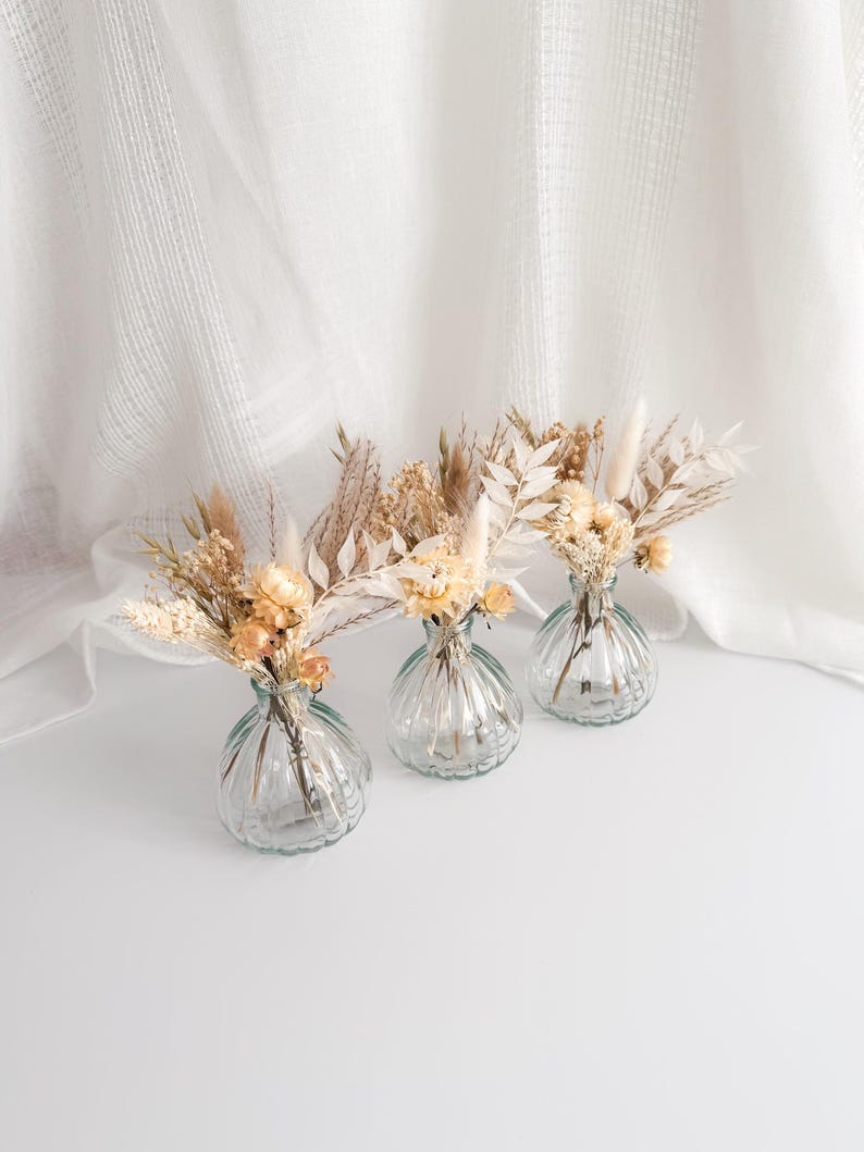 May include: Three small, clear glass vases with a ribbed design, each holding a delicate arrangement of dried flowers in shades of beige, cream, and light brown. The vases are arranged on a white surface, with a soft, white fabric backdrop.